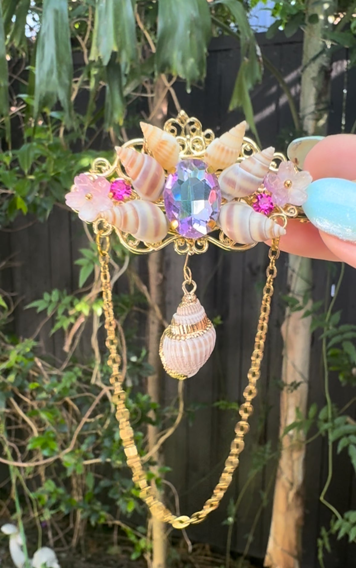 Decorative hair clip with shells and gemstones held against a natural background