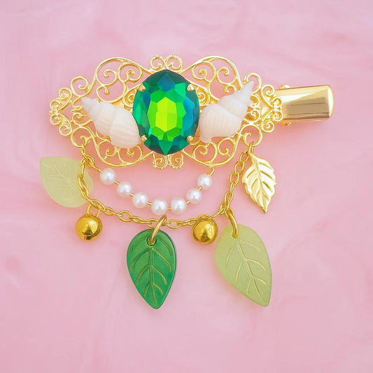 A gold-plated fantasy hair clip with a large green glass centerpiece, complemented by glass leaves and pearls, displayed against a pink background.