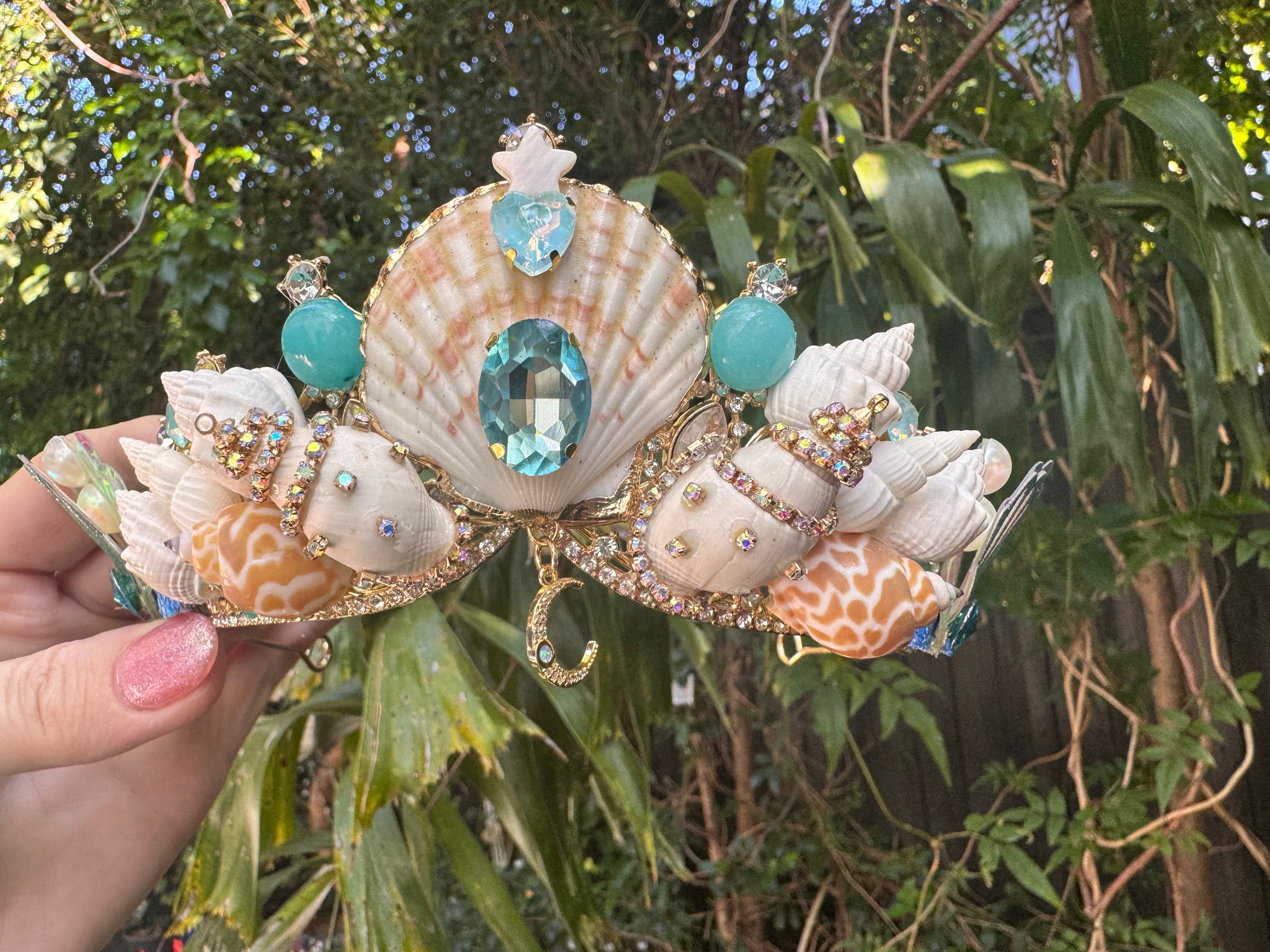 Seashell tiara with gemstones held by a hand against a green leafy background