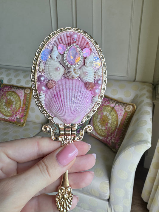 Decorative hand mirror with shell design held by a hand, against a neutral background.