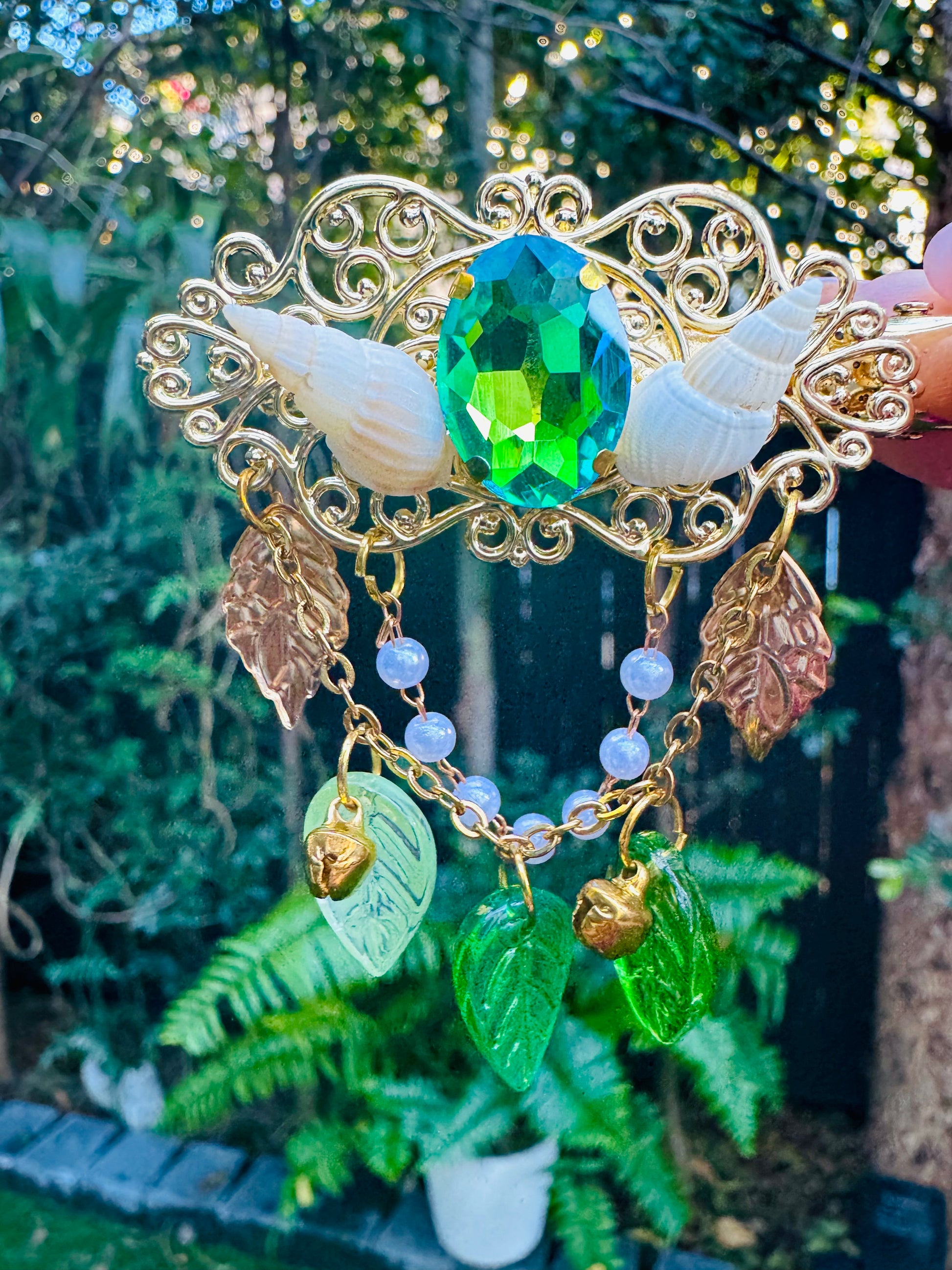 Ornate hair clip with green gemstone and leaf charms against a blurred natural background