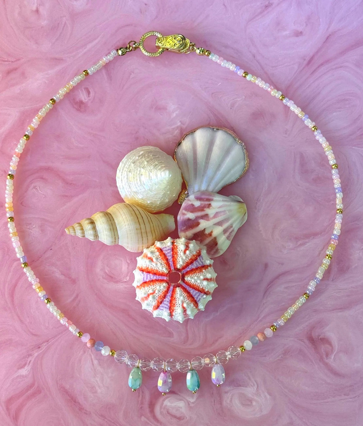 A beaded necklace with iridescent pastel beads, a gold clasp, and three dangling aqua and pale green teardrop charms, arranged on a pink marbled surface with decorative seashells.